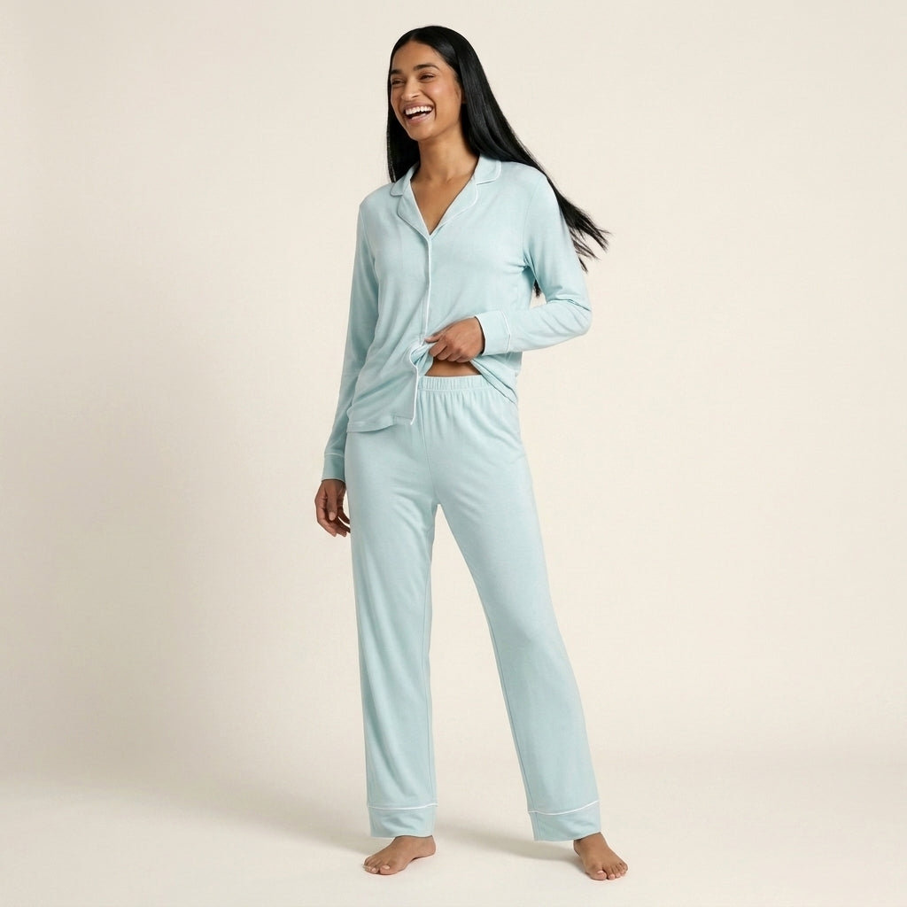 A woman modeling a light blue two-piece pajama set consisting of a long-sleeve button-front top and matching pants, photographed against a plain warm off-white studio background. Available in Ice Mint XS, Ice Mint S, Ice Mint M, Ice Mint L
