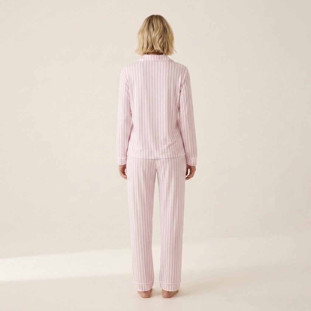 A woman viewed from behind wearing a matching two-piece pajama set with light pink and white vertical stripes, consisting of a long-sleeve top and straight-leg pants, against a plain cream studio background. Available in Pink Pinstripe XS, Pink Pinstripe S, Pink Pinstripe M, Pink Pinstripe L